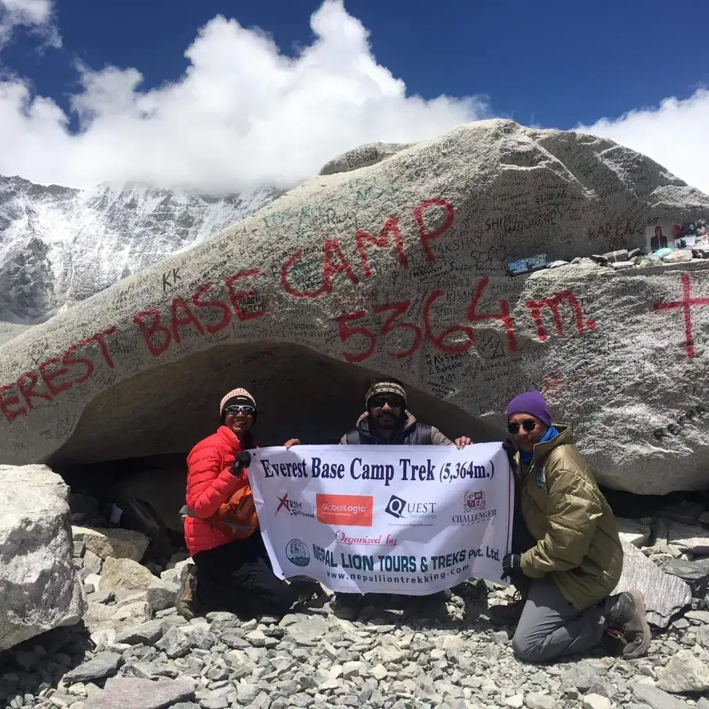 Three trekkers kneeling in front of the giant Everest Base Camp 5364m rock holding a banner from Nepal Lion Tours and Treks under a partly cloudy sky.