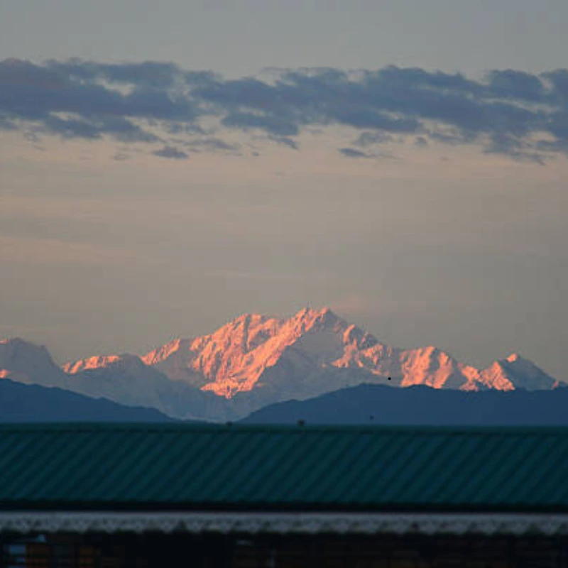 Pink sunset alpenglow illuminating high snow-covered Himalayan peaks across the distant horizon, viewed over a dark rooftop in the foreground.