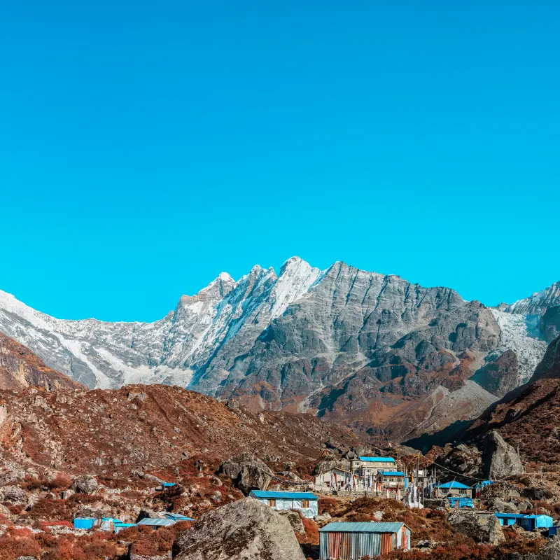Teahouses with bright blue and tin roofs nestled on a rugged hillside beneath a jagged snow-covered mountain.