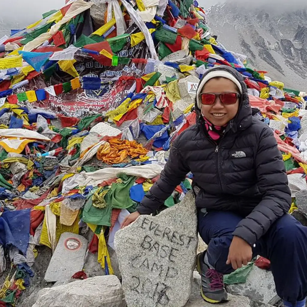A smiling female trekker in a black down jacket and sunglasses sitting in front of a colorful Buddhist prayer flag cairn and Everest Base Camp rock marker.