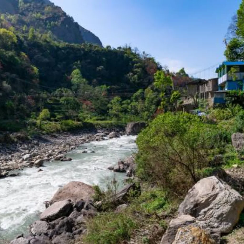 Rushing mountain river flanked by large boulders and green forested hills with rustic village houses and a suspension bridge along a trekking route in Nepal.