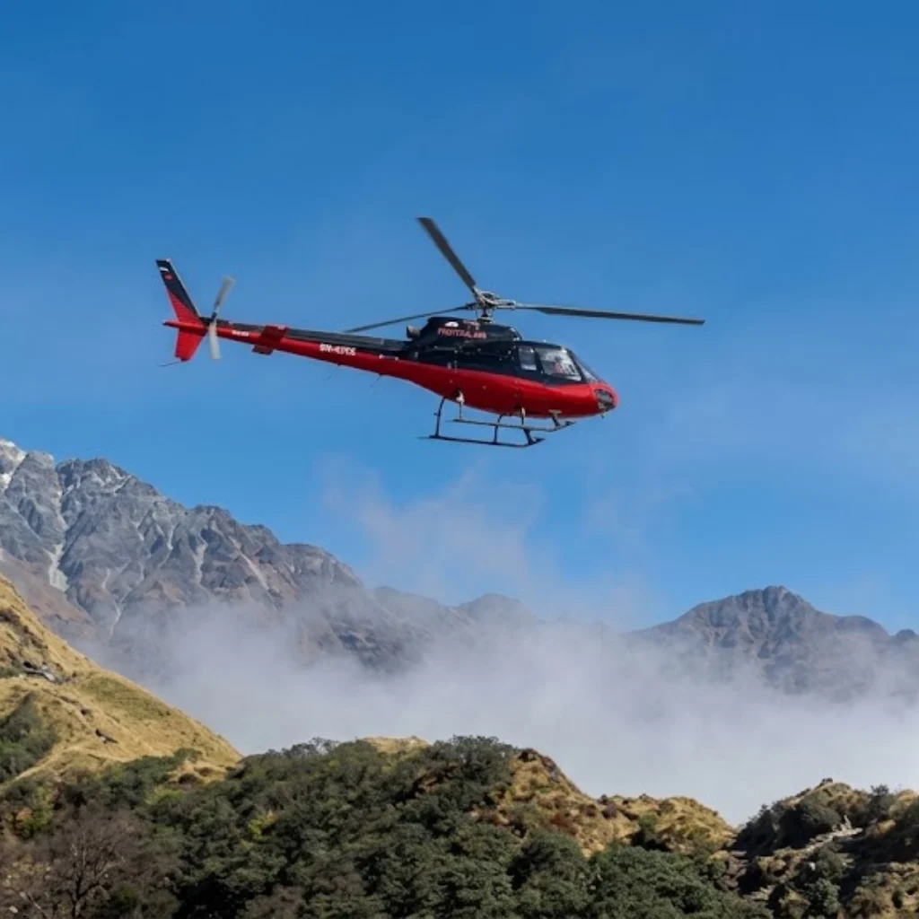 Red and black helicopter flying across clear blue skies over forested hills and misty mountain ridges.