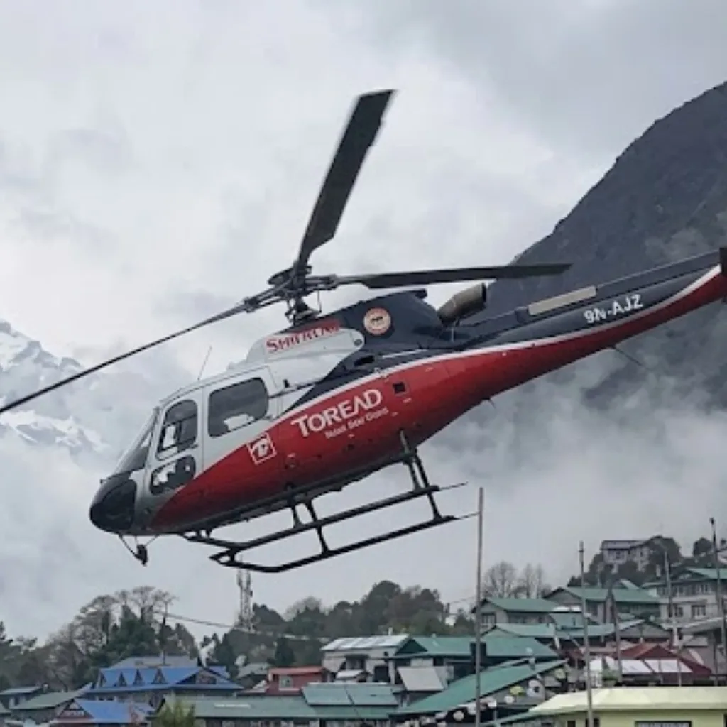 Red and white tour helicopter hovering above green-roofed lodges in a cloudy Himalayan mountain village.