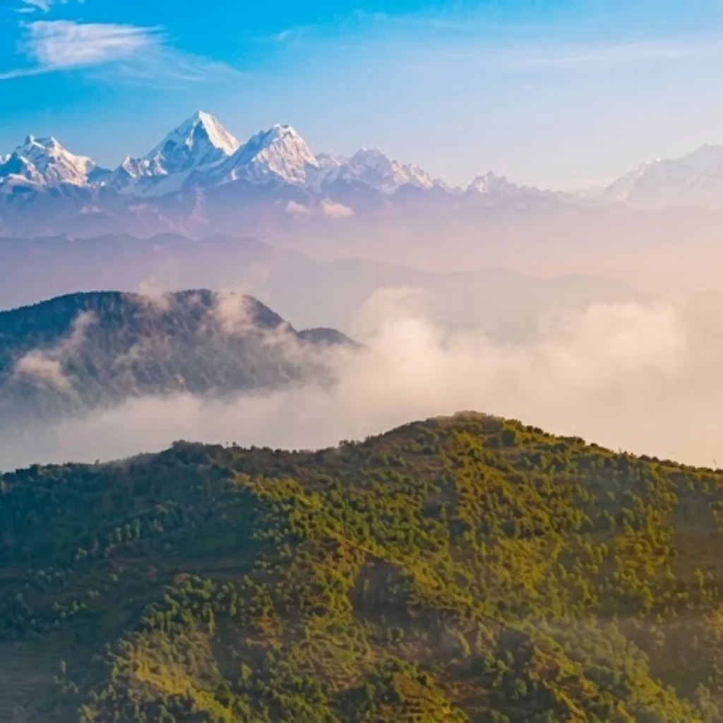 Sunlit green mountain ridges rising above soft morning clouds toward snow-capped Himalayan peaks.