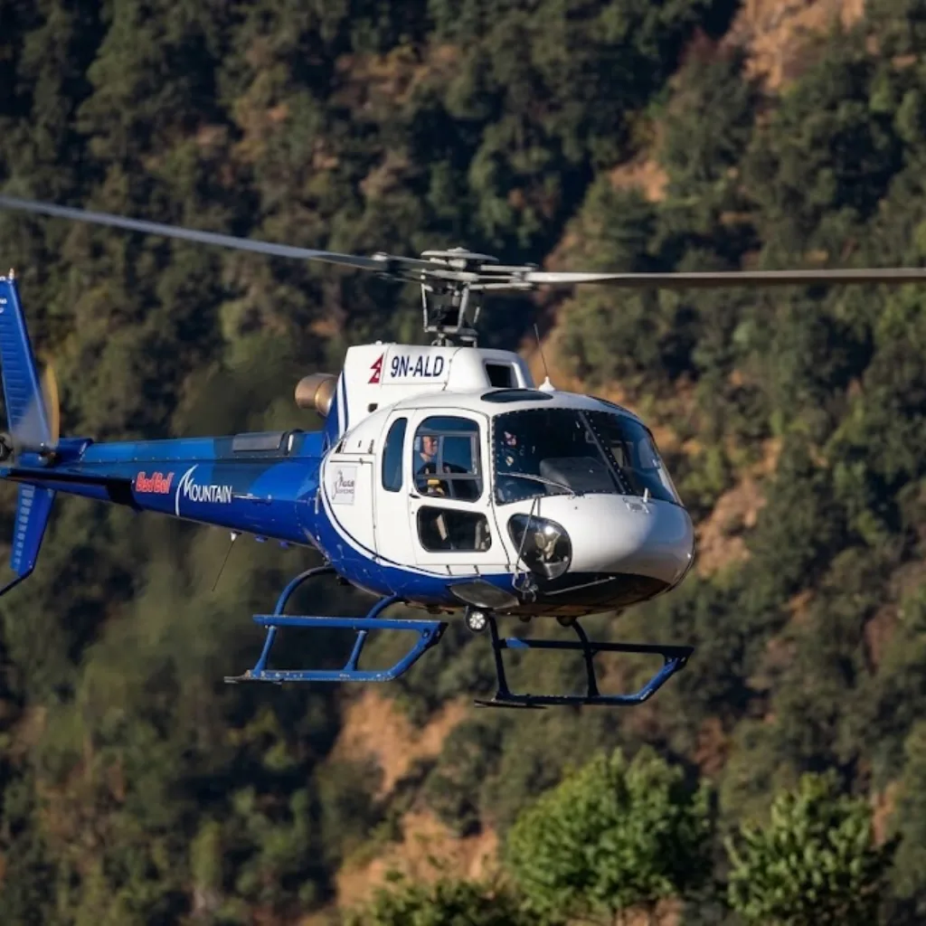 Blue and white charter helicopter flying low over lush green forested hills in the Himalayas of Nepal.