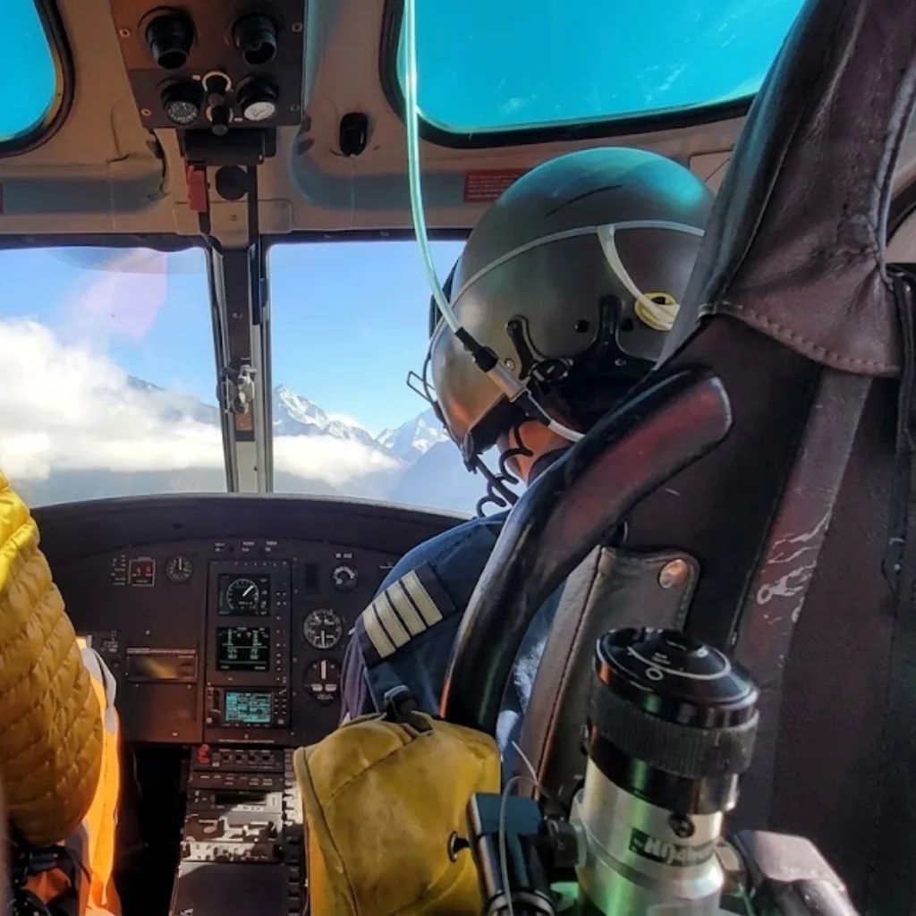View from inside a helicopter cockpit showing the pilot in a flight helmet flying toward snowy peaks.