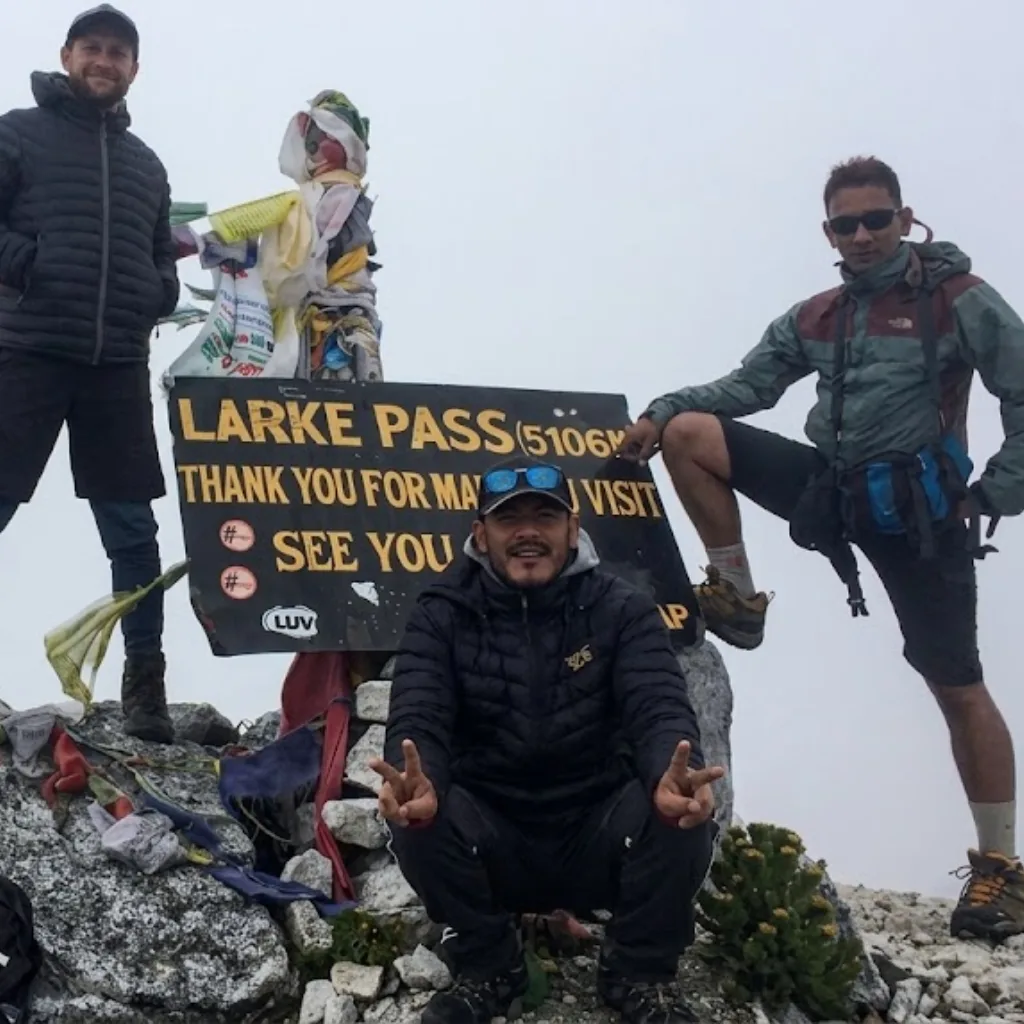 Three trekkers smiling and celebrating at the Larke Pass elevation sign at 5106 meters, draped in colorful Buddhist prayer flags on a foggy mountain peak.