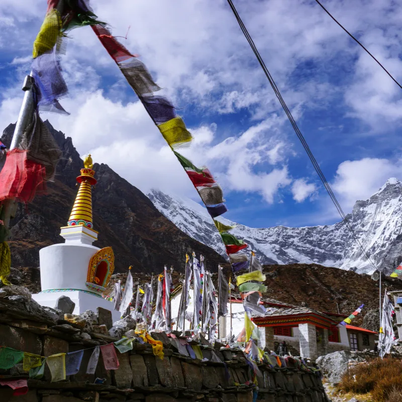 Traditional white Buddhist stupa draped with colorful prayer flags in front of towering snow-capped mountain peaks.
