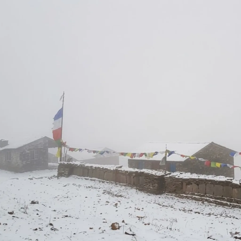 Stone mountain lodges and stone mani walls draped in Buddhist prayer flags during a dense, misty winter snowfall