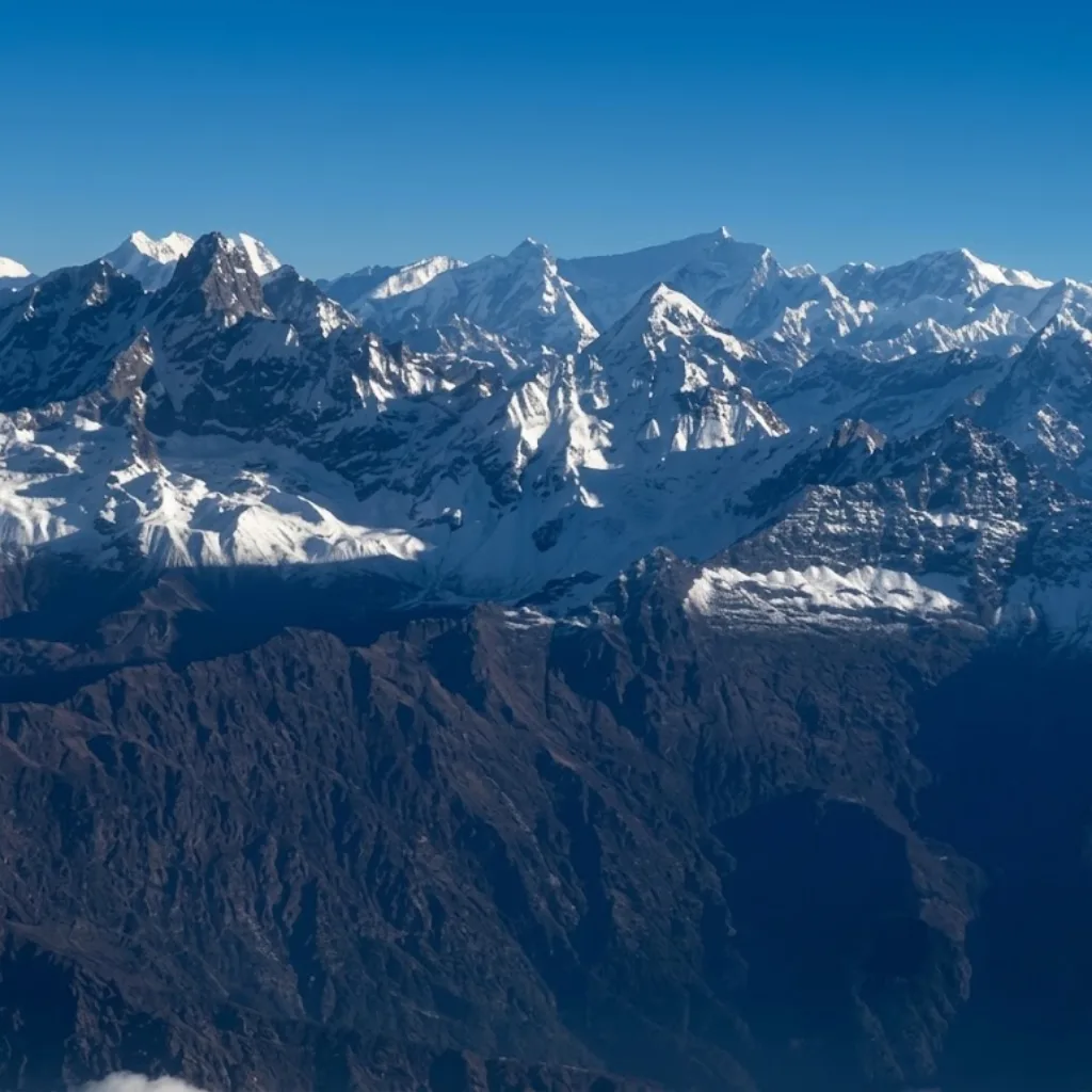 High-altitude aerial panorama showing sharp, snow-clad mountain peaks rising dramatically above dark rocky ridges and cloud layers.