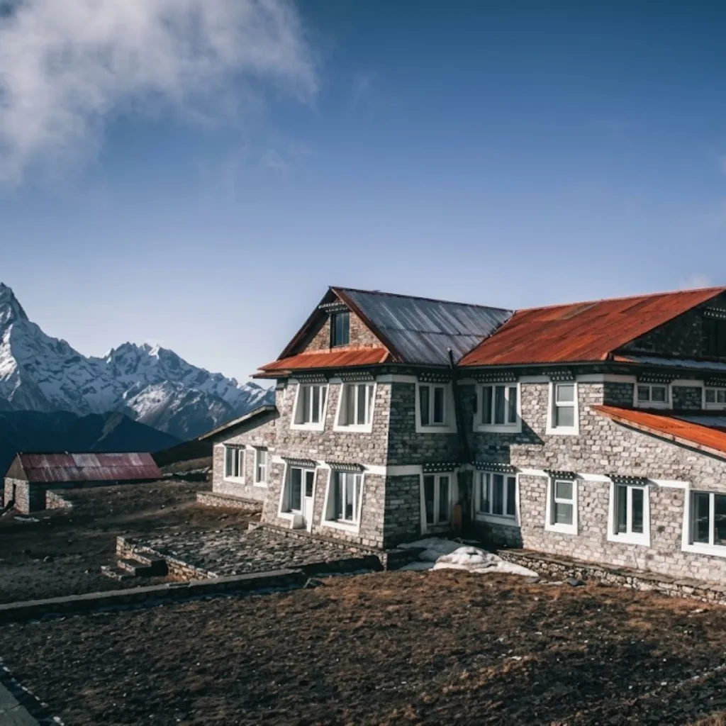 Stone mountain lodge with corrugated tin roof on an alpine plateau facing distant snowy Himalayan peaks.
