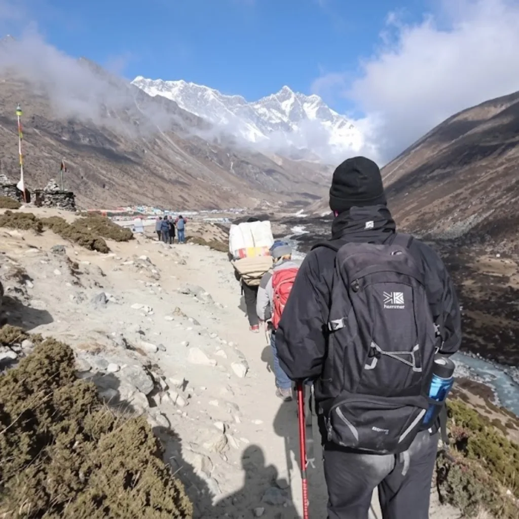 A group of trekkers and a porter walking along a dusty mountain path toward Mount Lhotse and snow-capped Himalayan peaks.