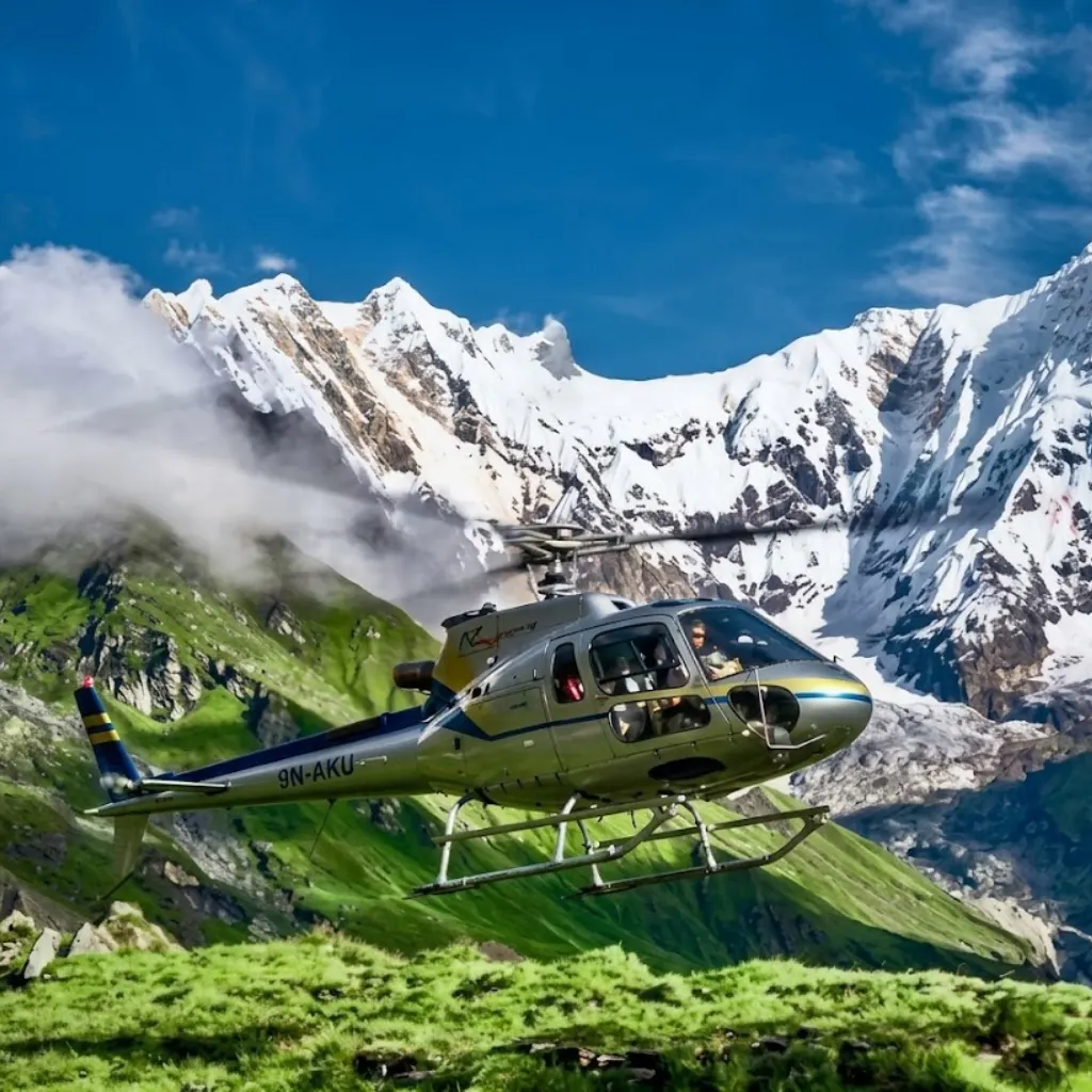 Silver helicopter flying over lush green grassy slopes before snow-capped Himalayan peaks and clouds.