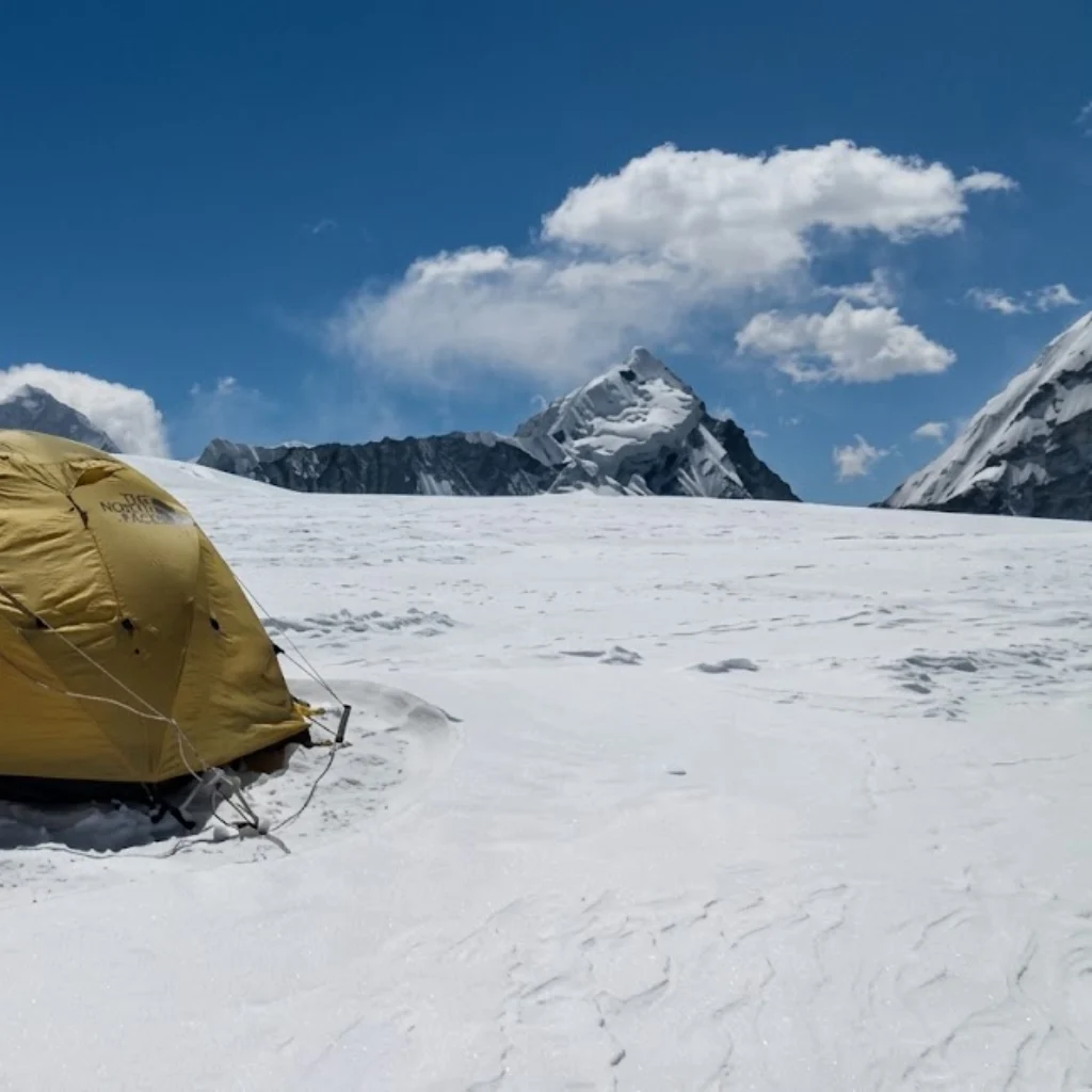 Yellow mountaineering dome tent pitched on a vast snowy glacier beneath high-altitude peaks and blue sky.