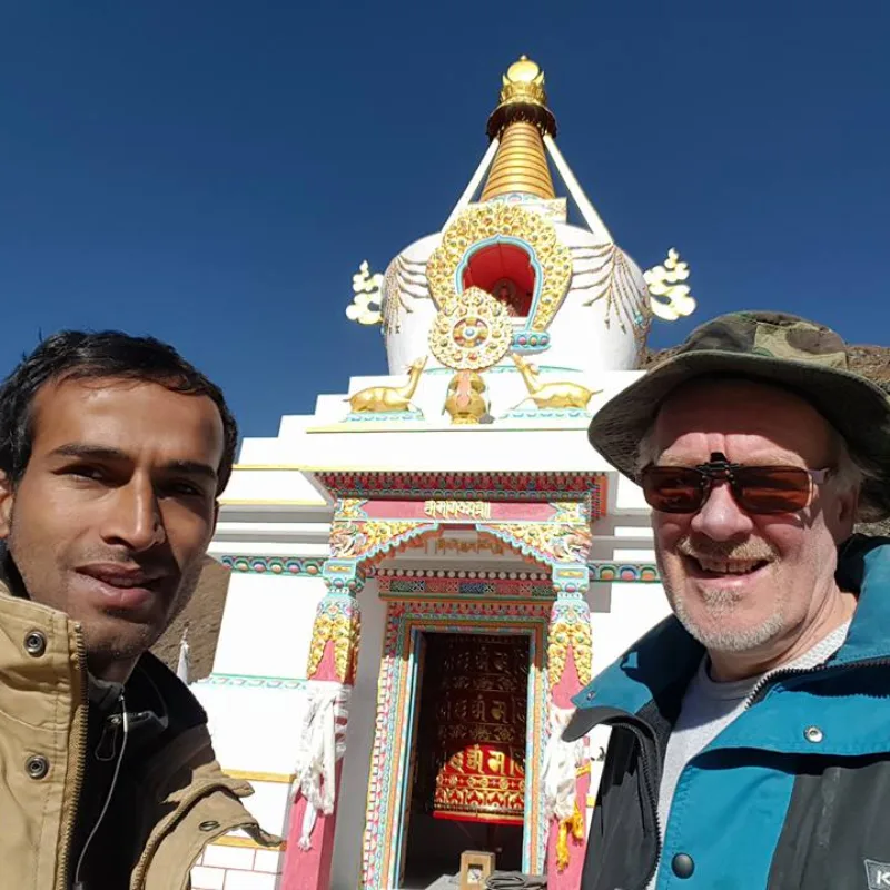 wo trekkers smiling for a selfie in front of a colorful Buddhist stupa with a prayer wheel under a clear blue sky.