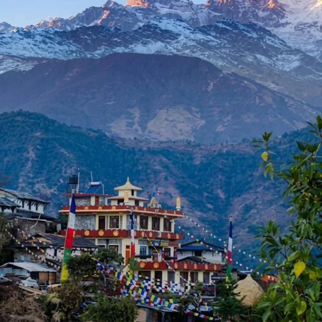 Tibetan Buddhist monastery with colorful prayer flags nestled on a hillside below snow-covered mountain walls.