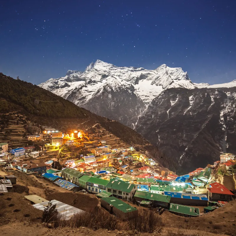 Horseshoe-shaped Sherpa town of Namche Bazaar illuminated at night beneath a starry sky with the massive snow-capped peak of Kongde Ri towering behind.