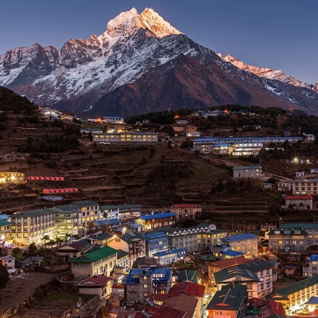 Evening view of illuminated lodges in Namche Bazaar beneath snow-dusted Himalayan peaks at dusk.