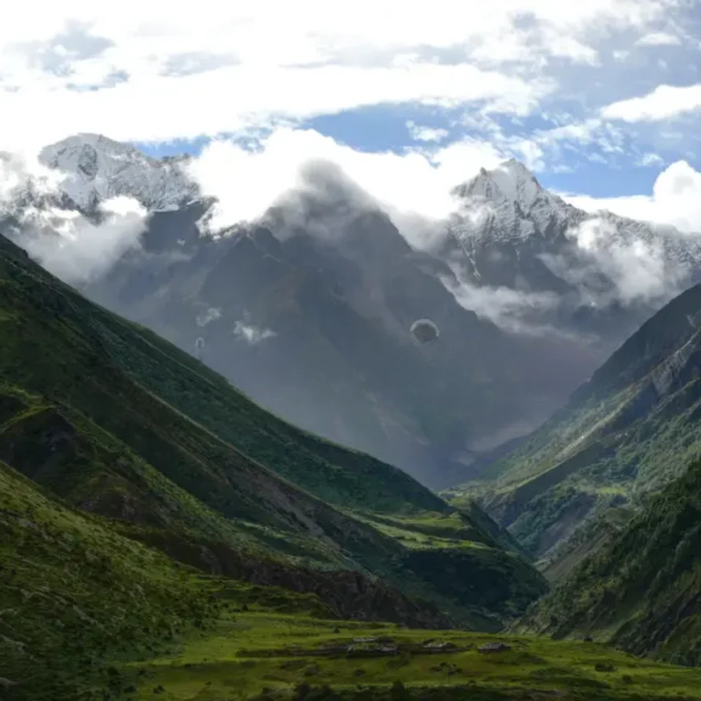 Lush green alpine valley floor leading into deep mountain gorges with mist and low clouds clinging to snow-dusted peaks.