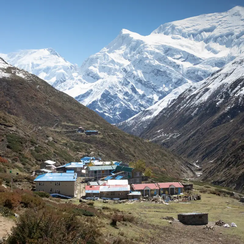 A cluster of mountain teahouses with bright blue and red roofs situated in a deep valley, surrounded by barren slopes and a massive snow-capped mountain.