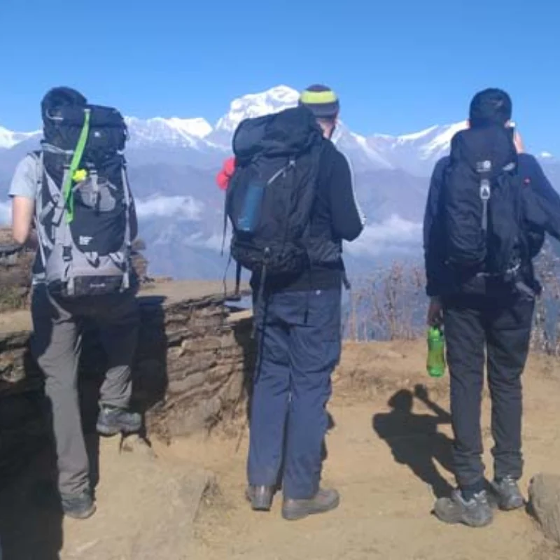 Three hikers with heavy backpacks standing on a stone ledge looking out at snow-covered Himalayan peaks.