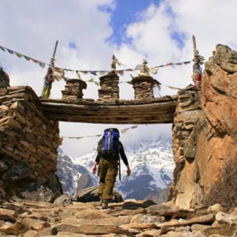 A trekker with a blue backpack walking through a traditional stone archway adorned with Buddhist prayer flags, heading toward snow-covered Himalayan mountains.
