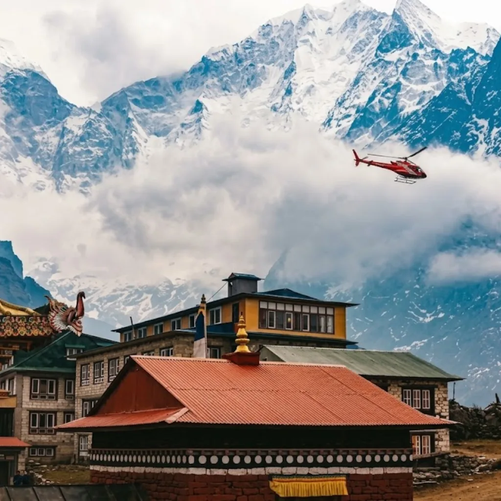 Red rescue helicopter flying over traditional Sherpa village lodges against towering, snow-covered mountains.