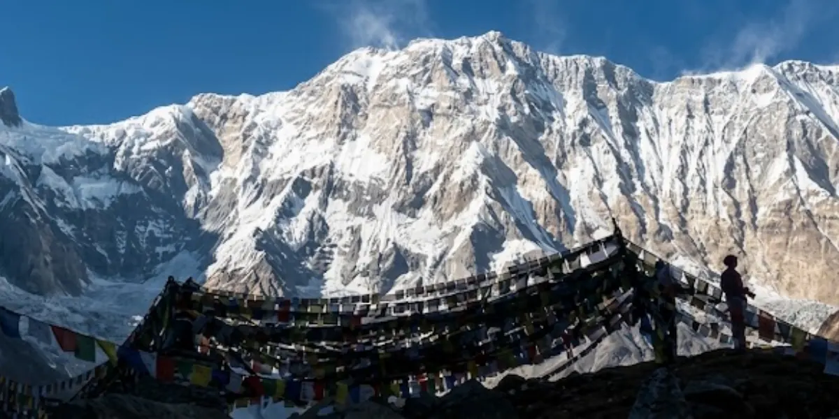 Silhouette of a person standing beside colorful Buddhist prayer flags overlooking the massive south face of Mount Annapurna.